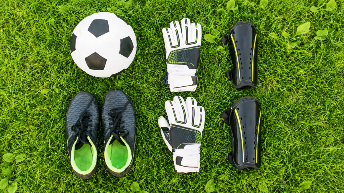 Materiais de futebol com bola, chuteiras, luvas de goleiro e caneleiras sobre a grama.