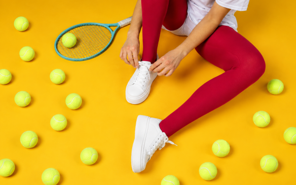 Pessoa sentada amarrando tênis branco, com raquete e bolas de tênis sobre fundo amarelo